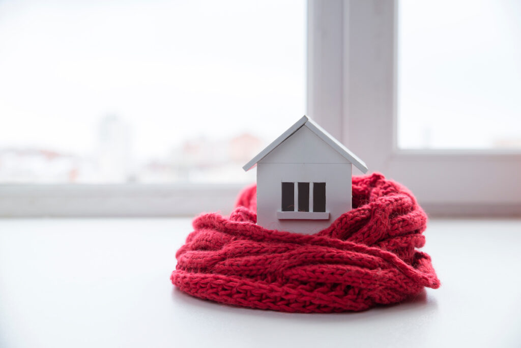 house in winter - heating system concept and cold snowy weather with model of a house wearing a knitted cap.
